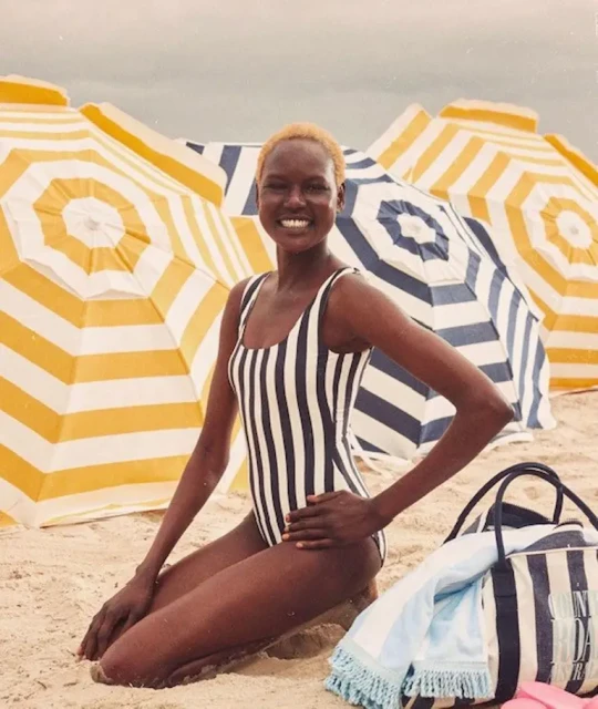 A tanned woman smiles while kneeling on a sandy beach in a striped swimsuit, next to a beach bag and striped umbrellas.