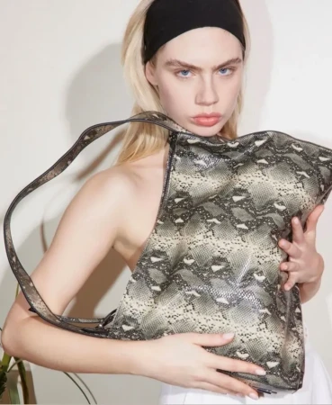 Young woman with blond hair poses indoors, holding a snakeskin print purse with a shoulder strap in a close up shot.