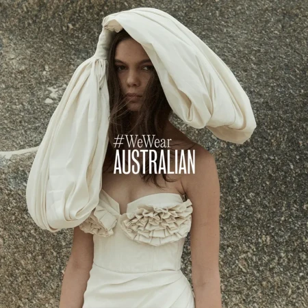 Model wearing a structured cream dress and sculptural headpiece against a rocky textured outdoor background with campaign text.