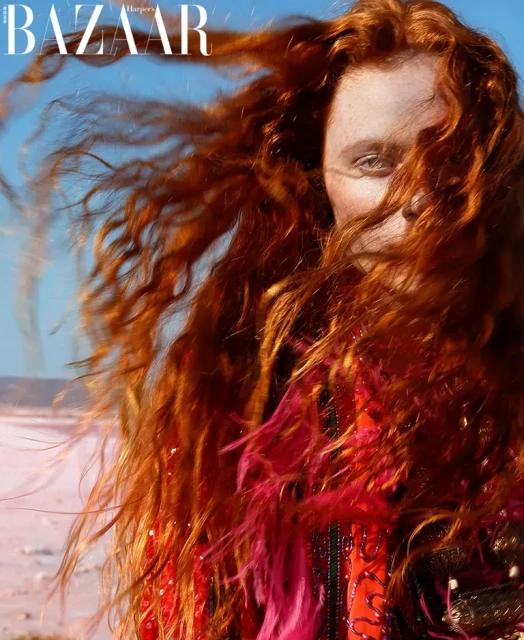 Fashion editorial portrait of a young adult woman with long red hair wearing a red patterned jacket outdoors in a desert setting.