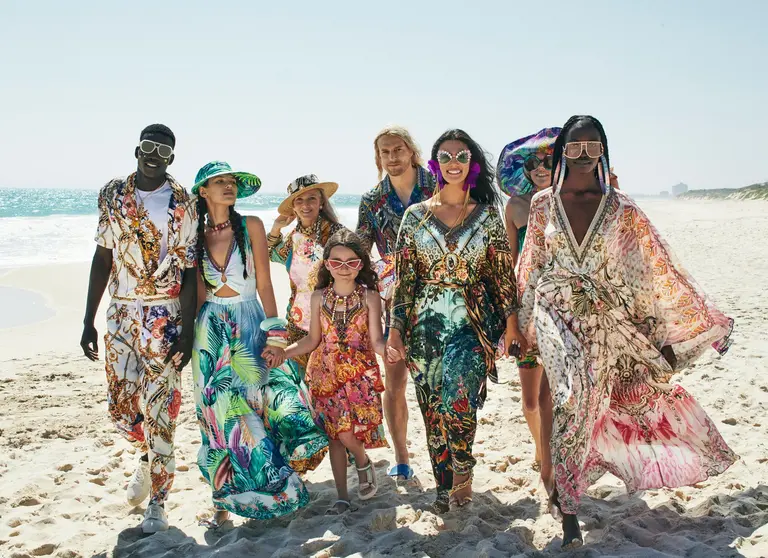 Group of diverse models pose on a sunny beach wearing colorful patterned resort wear, sunglasses, and hats in a fashion editorial.