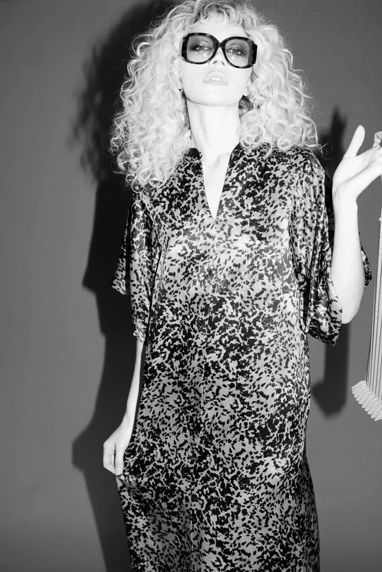 Woman with blonde curly hair and oversized sunglasses wearing a patterned dress indoors against a solid background in a studio-style setting.