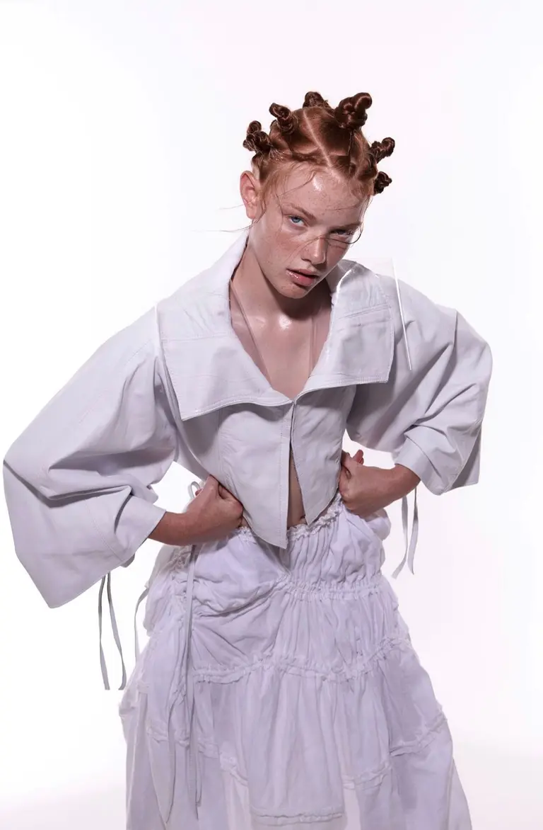 Young adult female fashion model in white crop top and skirt with her hands on her waist posing in a photo studio against a white background.