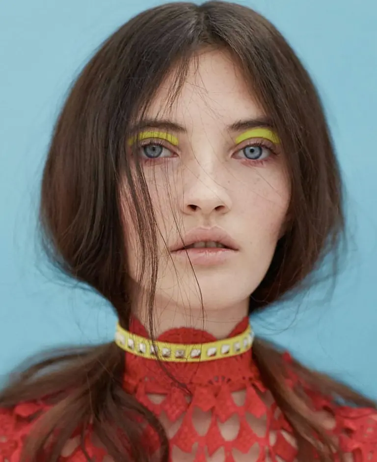 Portrait of a brunette woman with yellow eyeshadow and a red patterned top against a light blue background in a beauty studio shot.