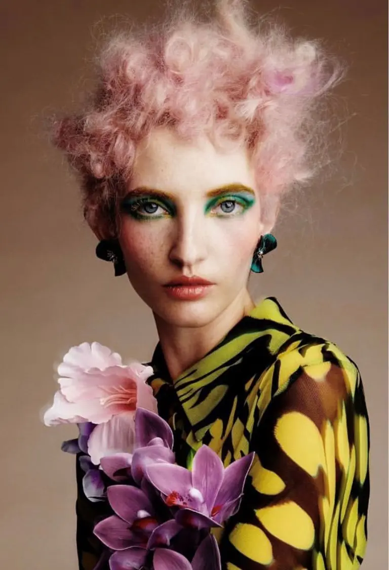 Close-up of a stylish young woman with creative green eyeshadow, pink fluffy hair, and floral accents. She wears a patterned yellow blouse.