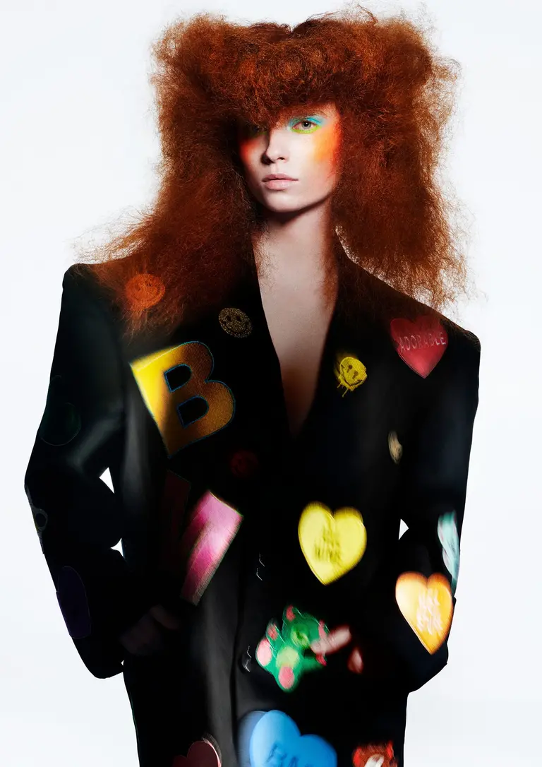 Fashion photo features a woman with voluminous red hair wearing a black blazer decorated with colorful heart and letter-shaped pins.