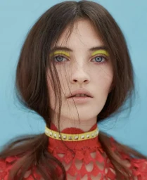 Close-up portrait of a woman with vibrant yellow eyeshadow, a studded yellow choker, and a red patterned top against a pale blue background.