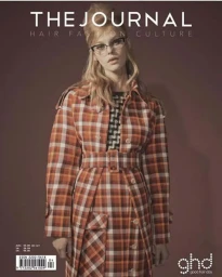 Model wearing a plaid belted coat and cat-eye glasses with voluminous retro hairstyle against a dark background