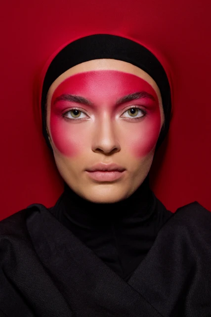 Close-up studio portrait of a woman with vibrant red face paint and a black head covering, set against a matching red backdrop.