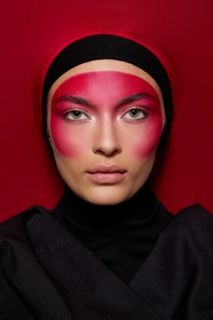 Close-up studio portrait of a woman with vibrant red face paint and a black head covering, set against a matching red backdrop.