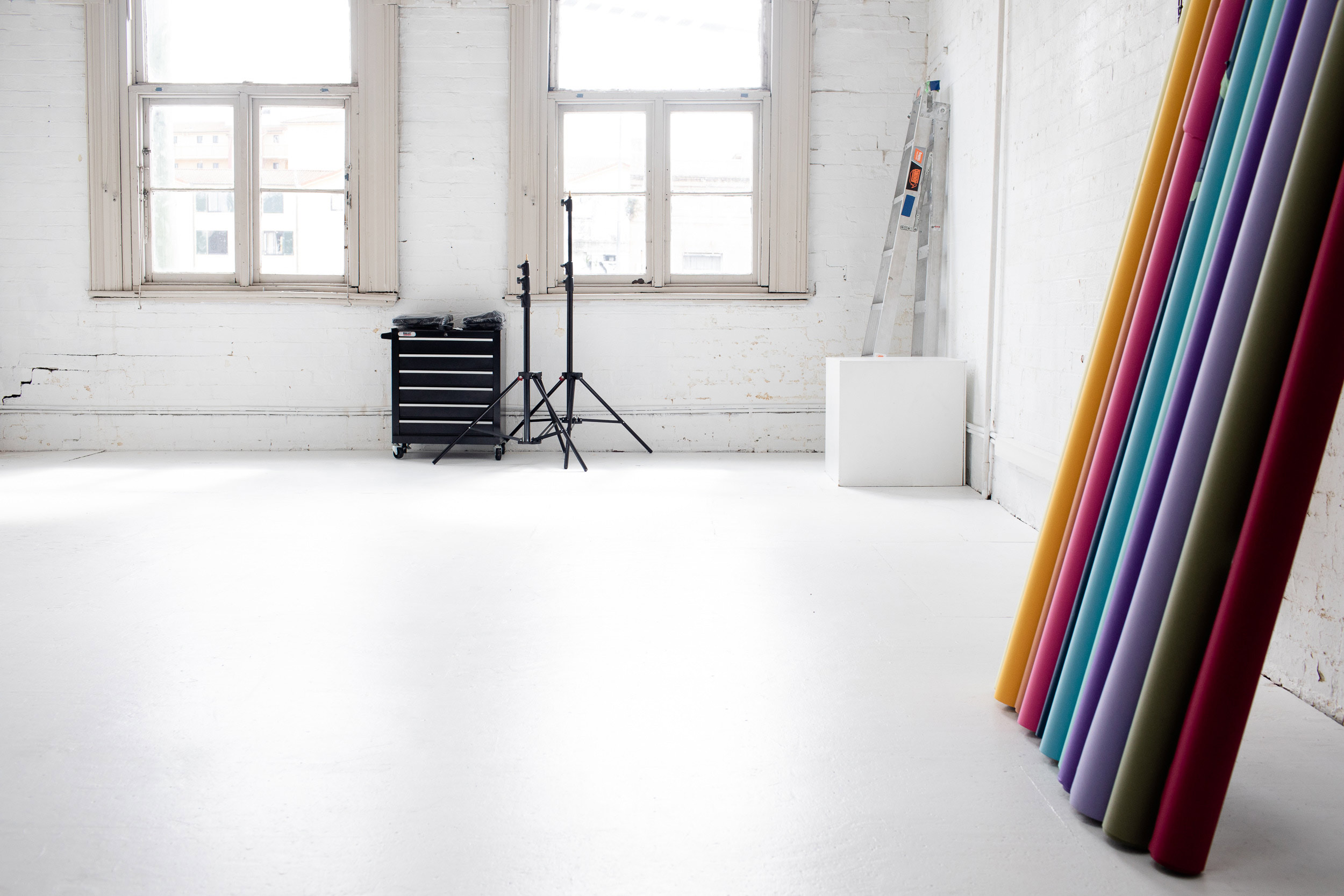 Bright photography studio interior with white walls and floor, natural light from windows, and colorful backdrop rolls.