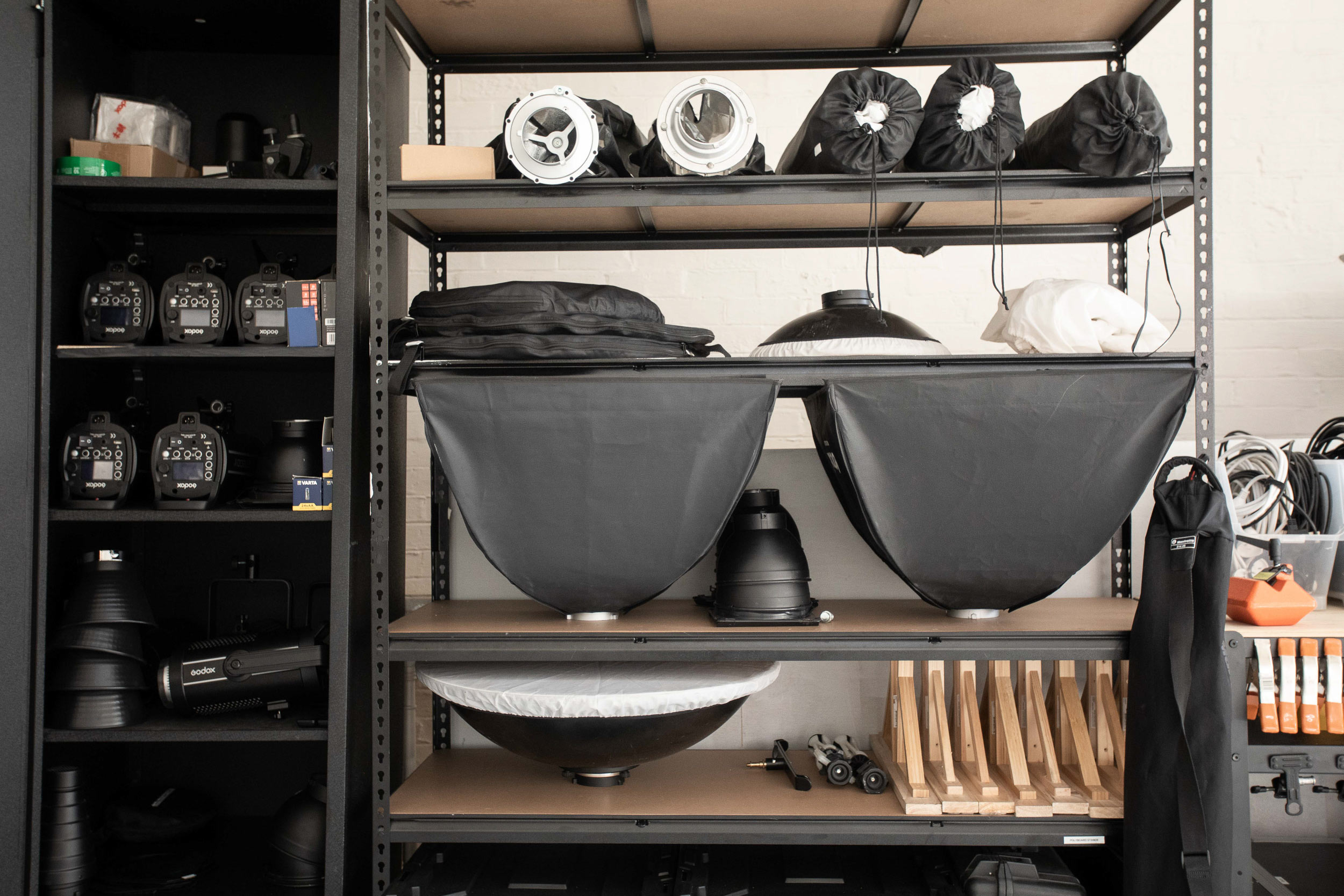 Photo studio equipment organized on metal shelving and in a cabinet, including softboxes, reflectors, and lighting control units.