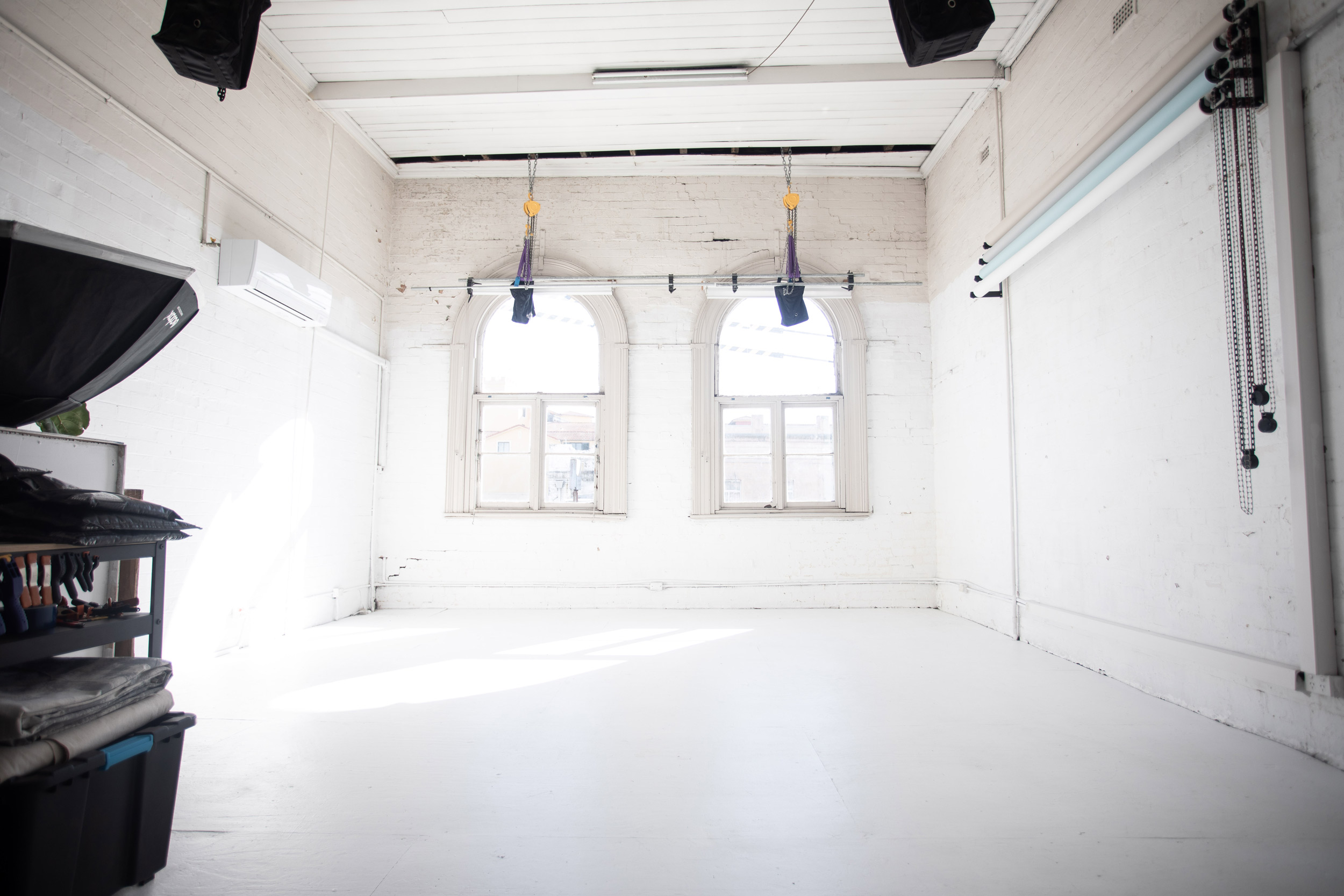 Bright, empty photo studio featuring white walls, two arched windows, and visible lighting equipment for professional photography.