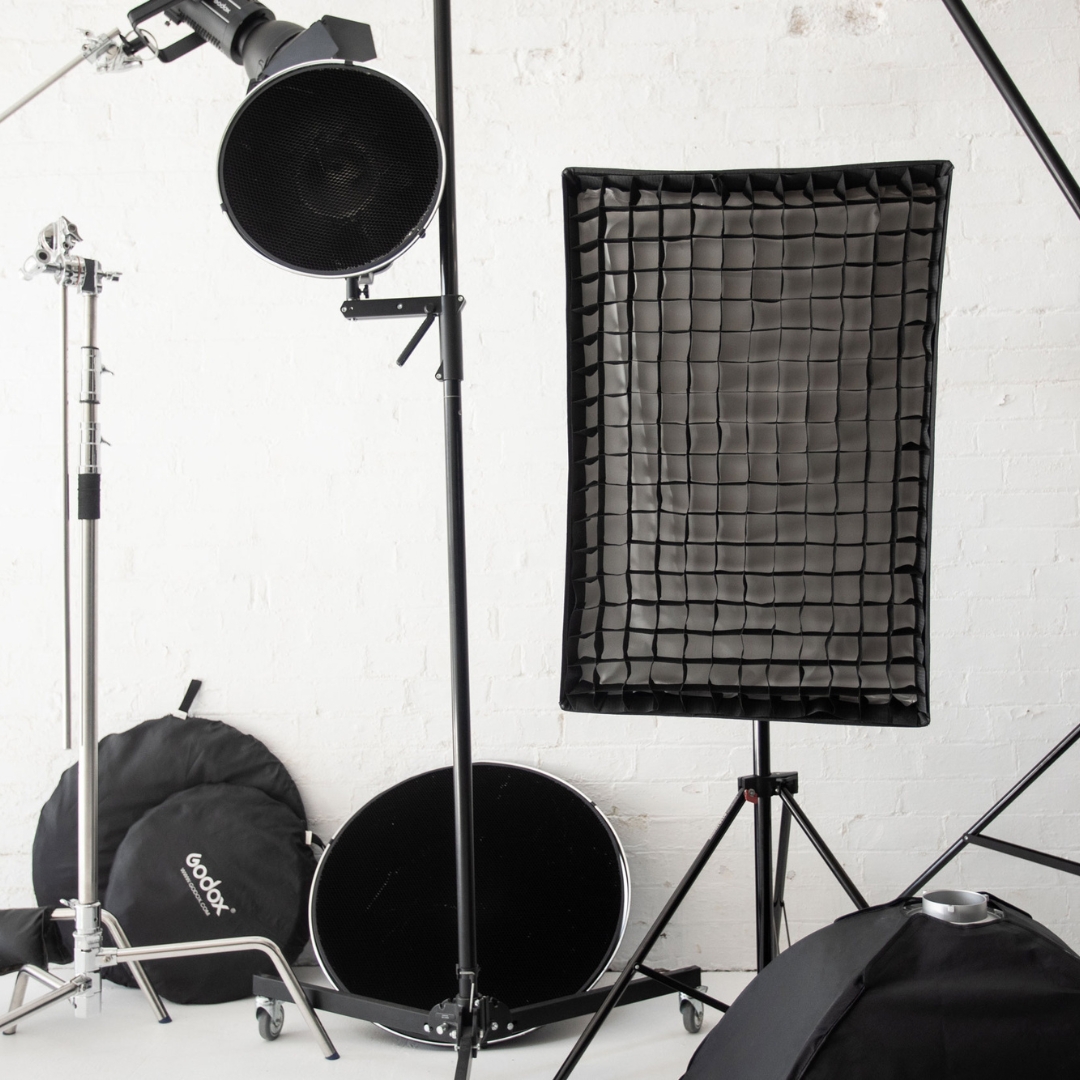 Photo studio equipment: softbox, light stands, reflector, and umbrella against a white brick wall in a photography studio setup.