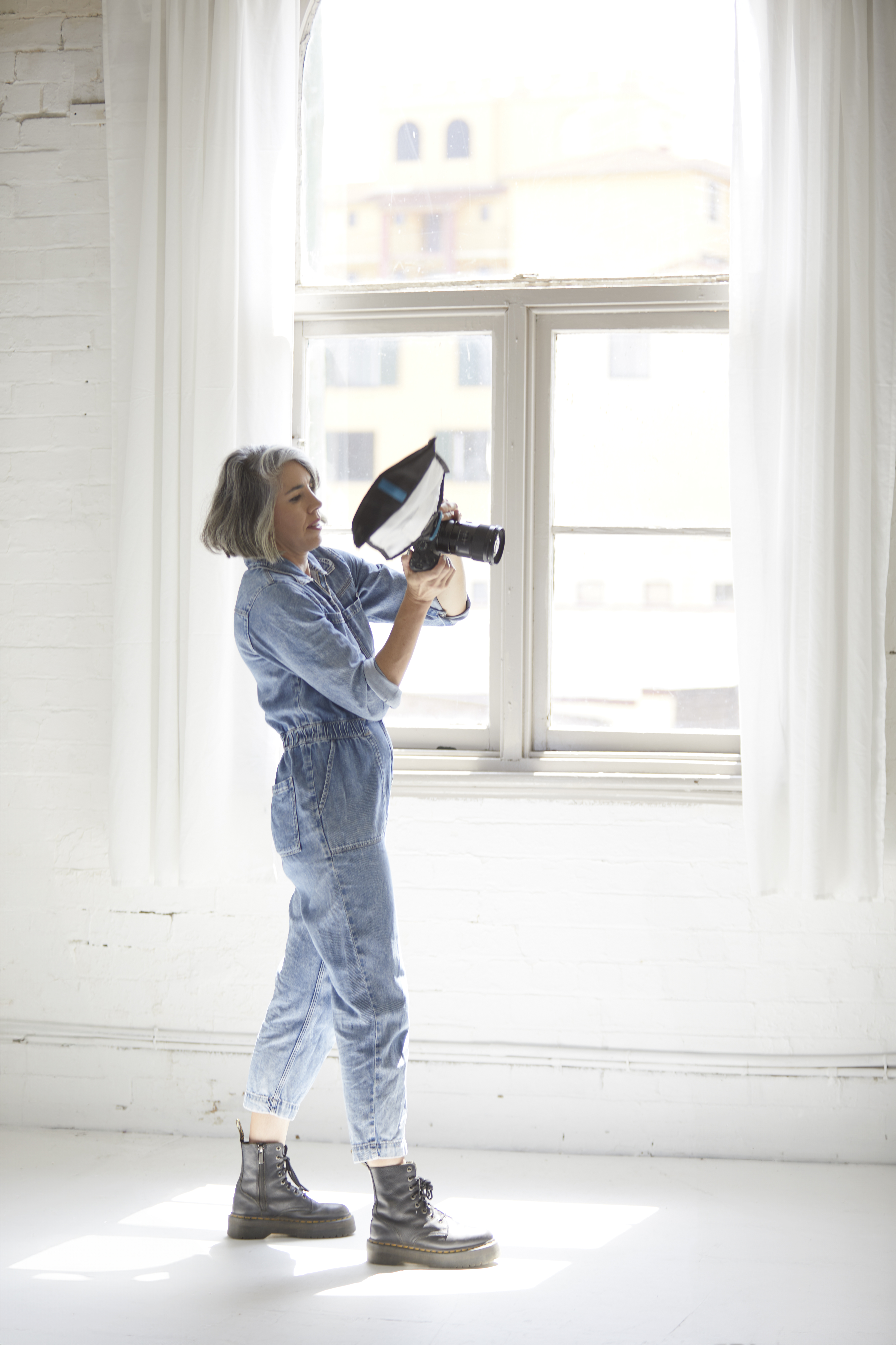 Adult woman in denim jumpsuit takes a photo with a digital camera and speedlight in a bright white photo studio with natural light.