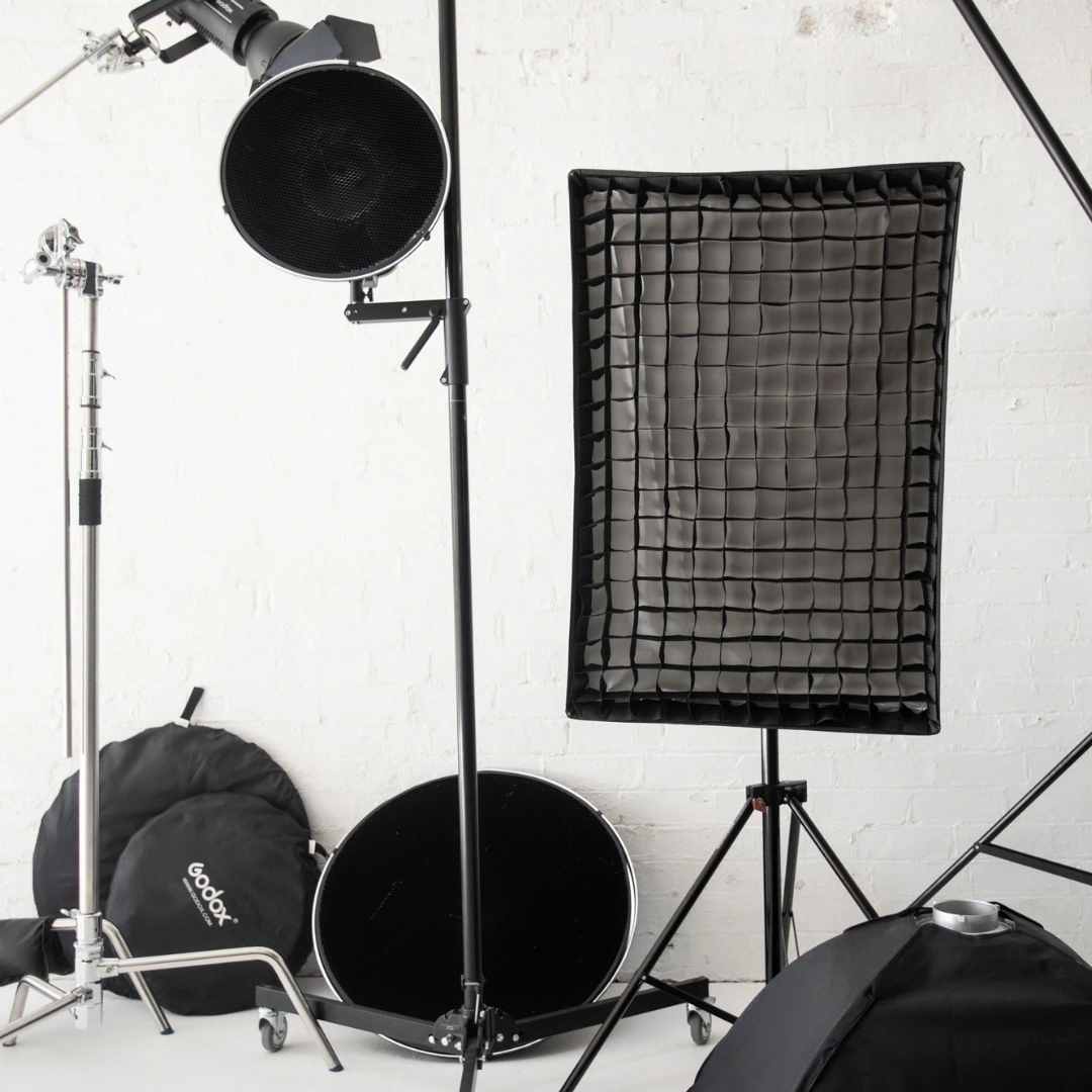 Professional photography studio equipment including studio lights, softboxes, stands, and reflectors against a white brick wall.