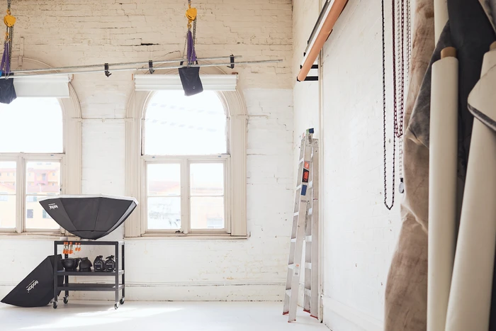 Spacious photo studio interior featuring arched windows, white brick walls, a metal ladder, and studio lighting equipment.