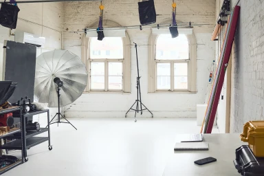 Professional photography studio interior featuring arched windows, white brick walls, lighting equipment, and a laptop on a desk