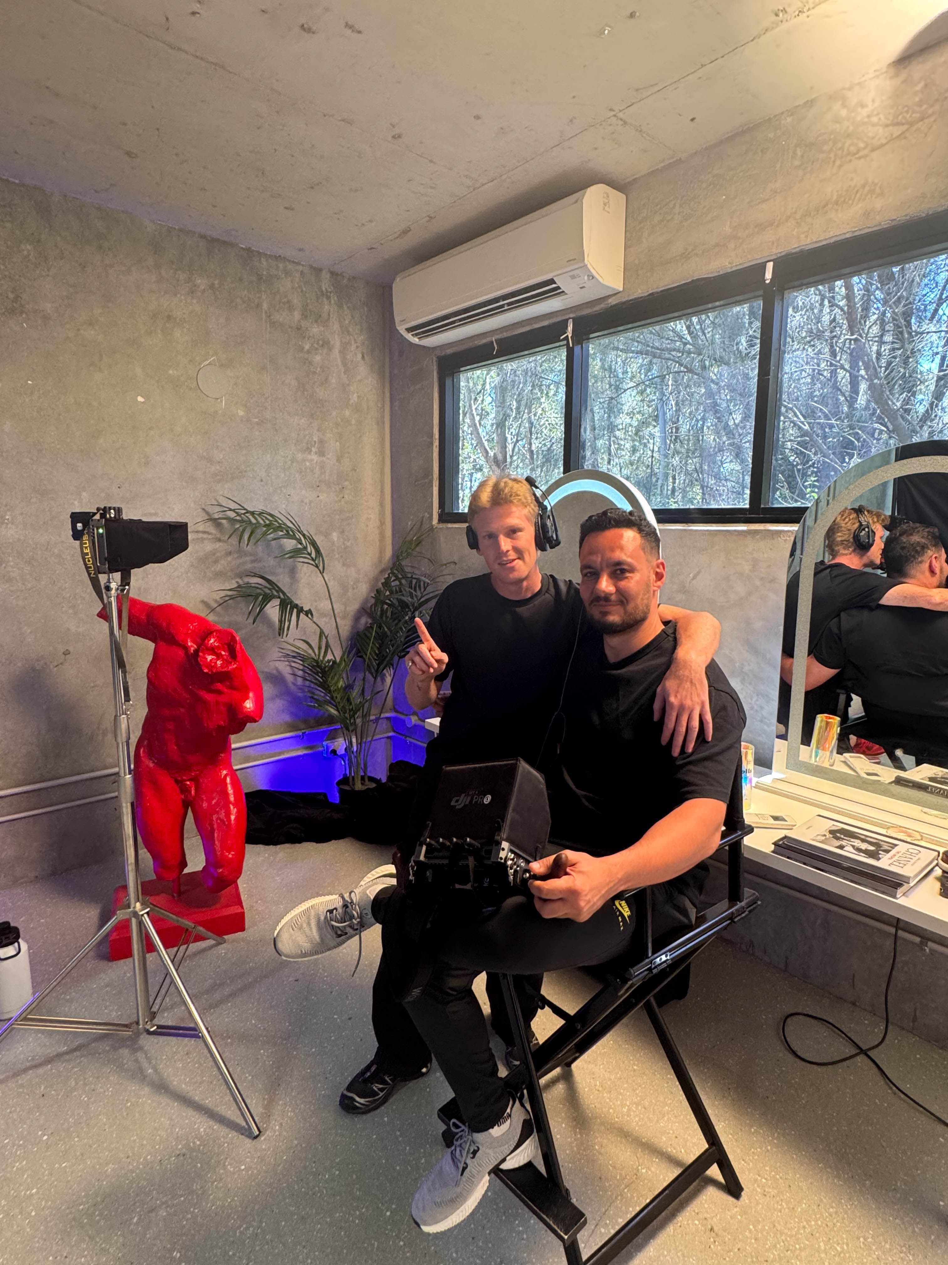 Two adult men pose indoors; one is seated in a director's chair, holding film equipment, and the other is standing with arm around him.