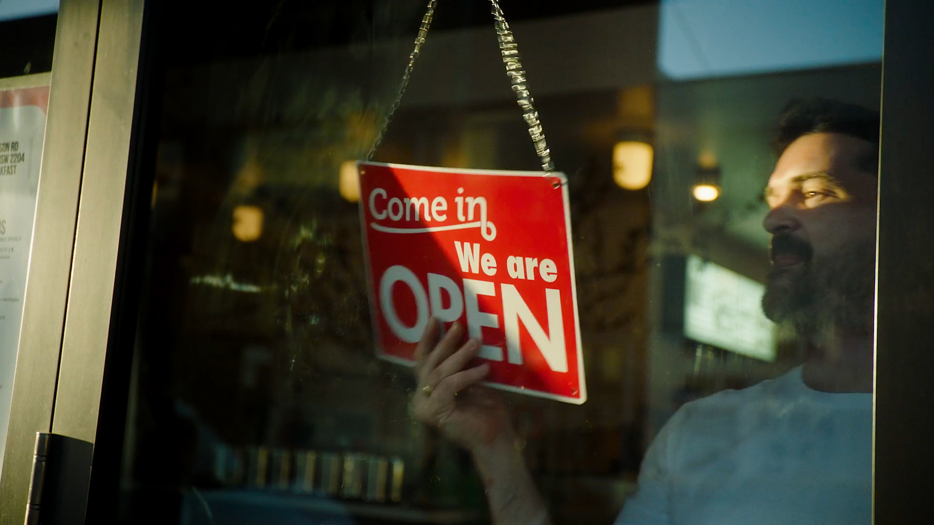 A man inside a restaurant is holding an 'Open' sign with a red background and white letters, indicating the business is open.