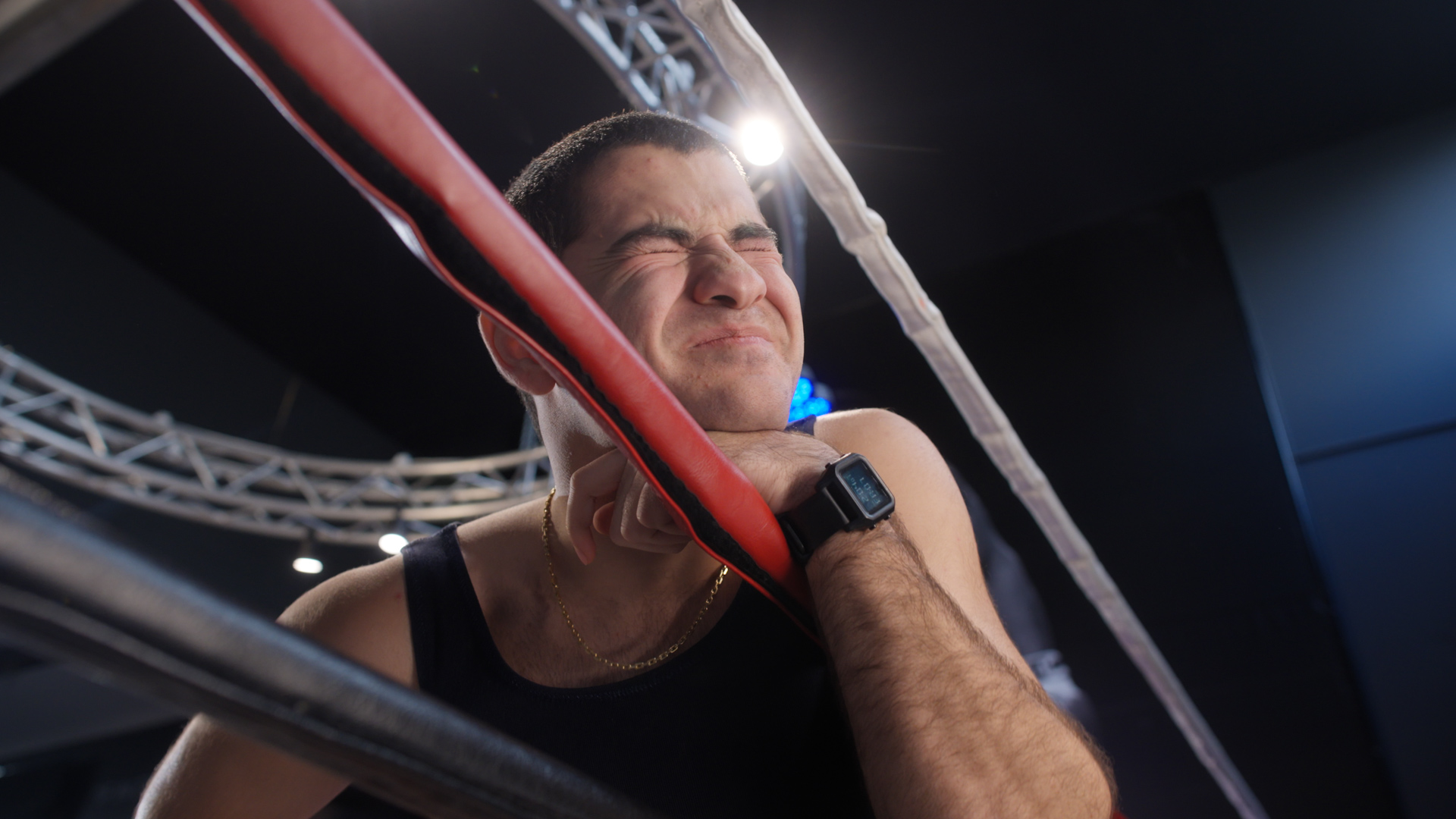 Male athlete at a boxing ring with face contorted near red corner. View from below looking up with bright overhead lighting.