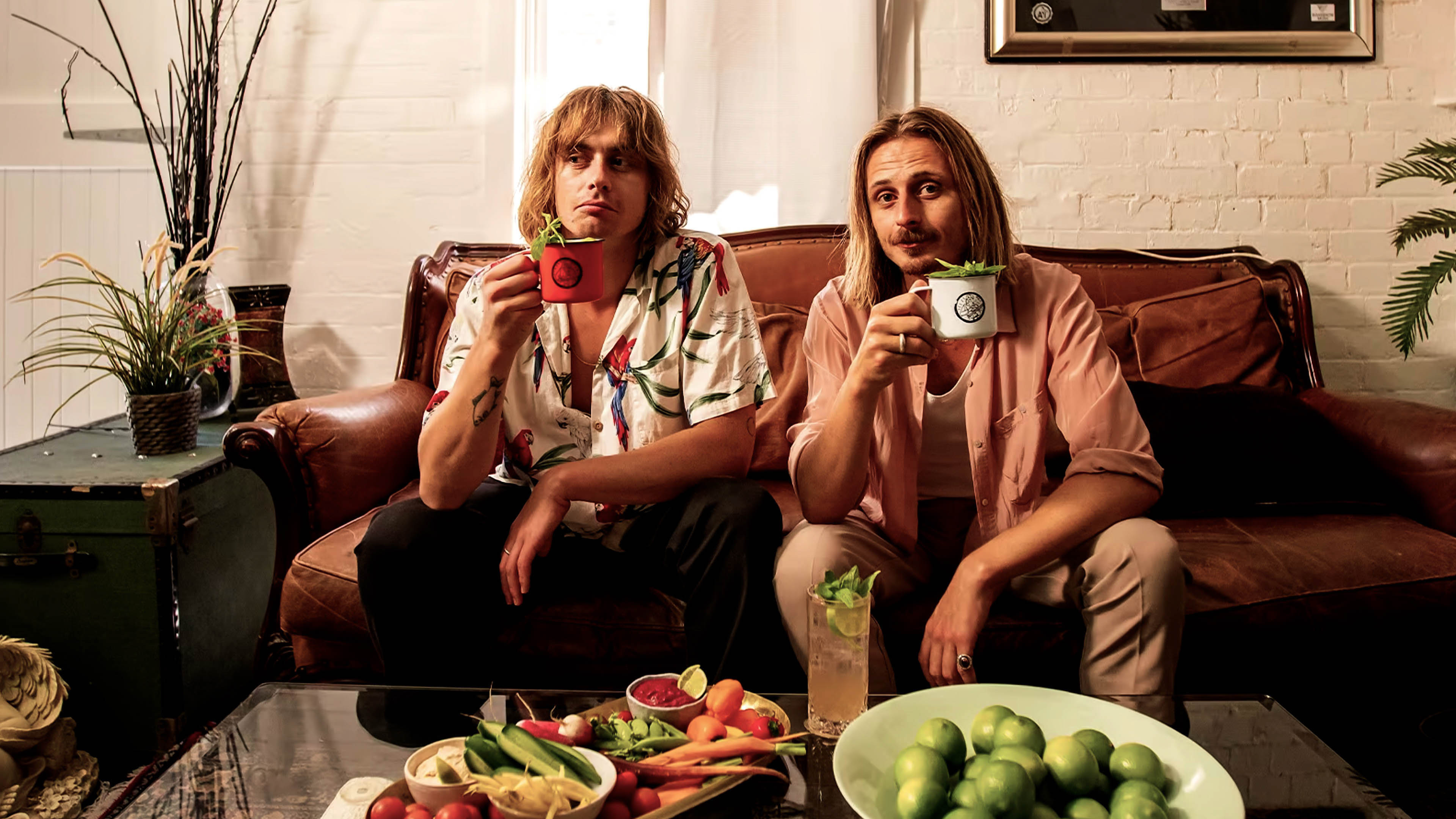 Two men sit on a couch, holding mugs, with snacks on a glass table in front of them in a relaxed indoor setting.