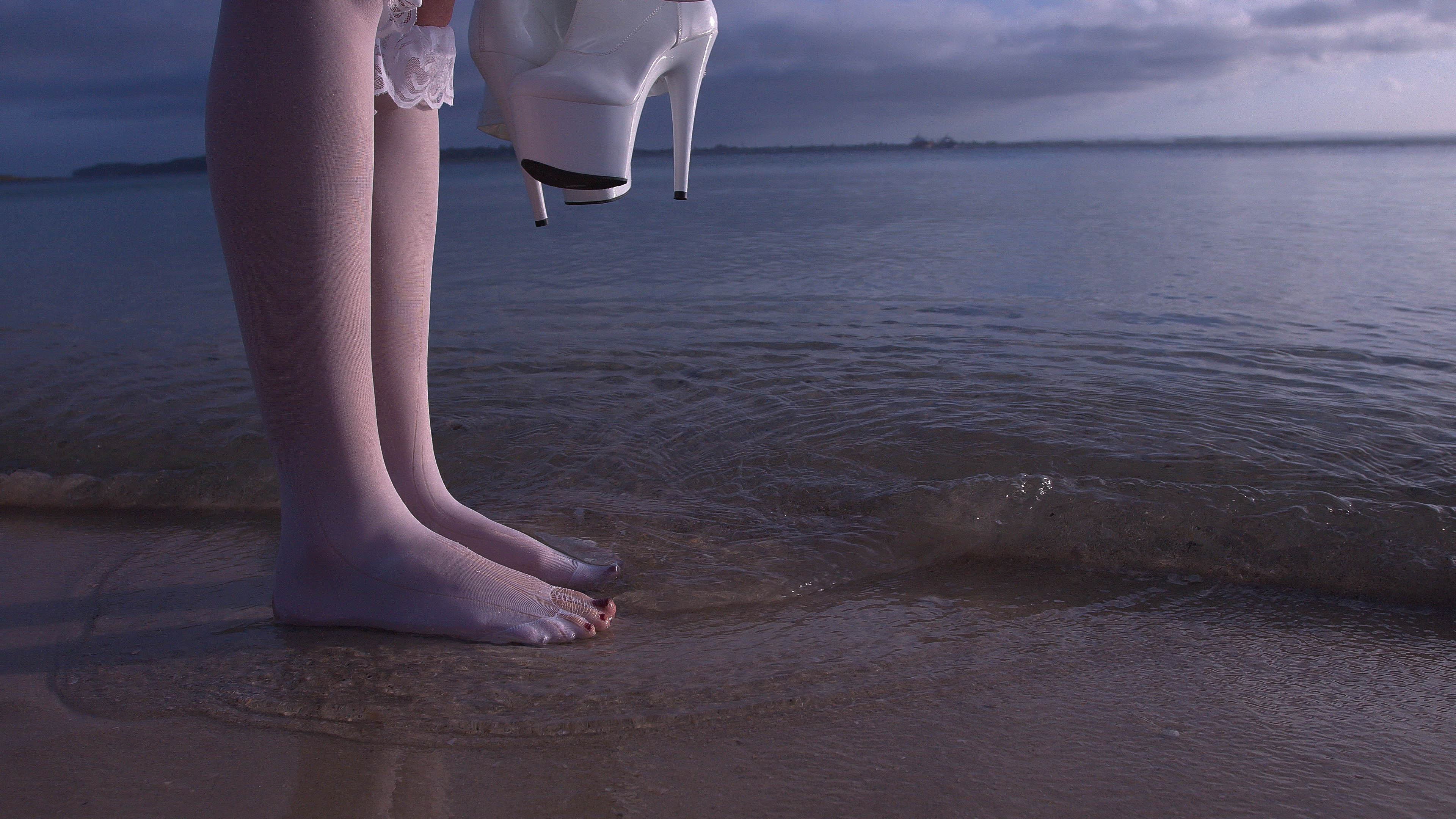 Legs of a woman wearing white stockings standing in ocean surf holding platform heels, with a cloudy sky and water background.