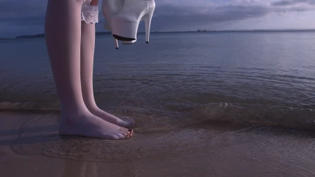 Legs of a person wearing white tights and holding high heels standing in shallow water at the beach