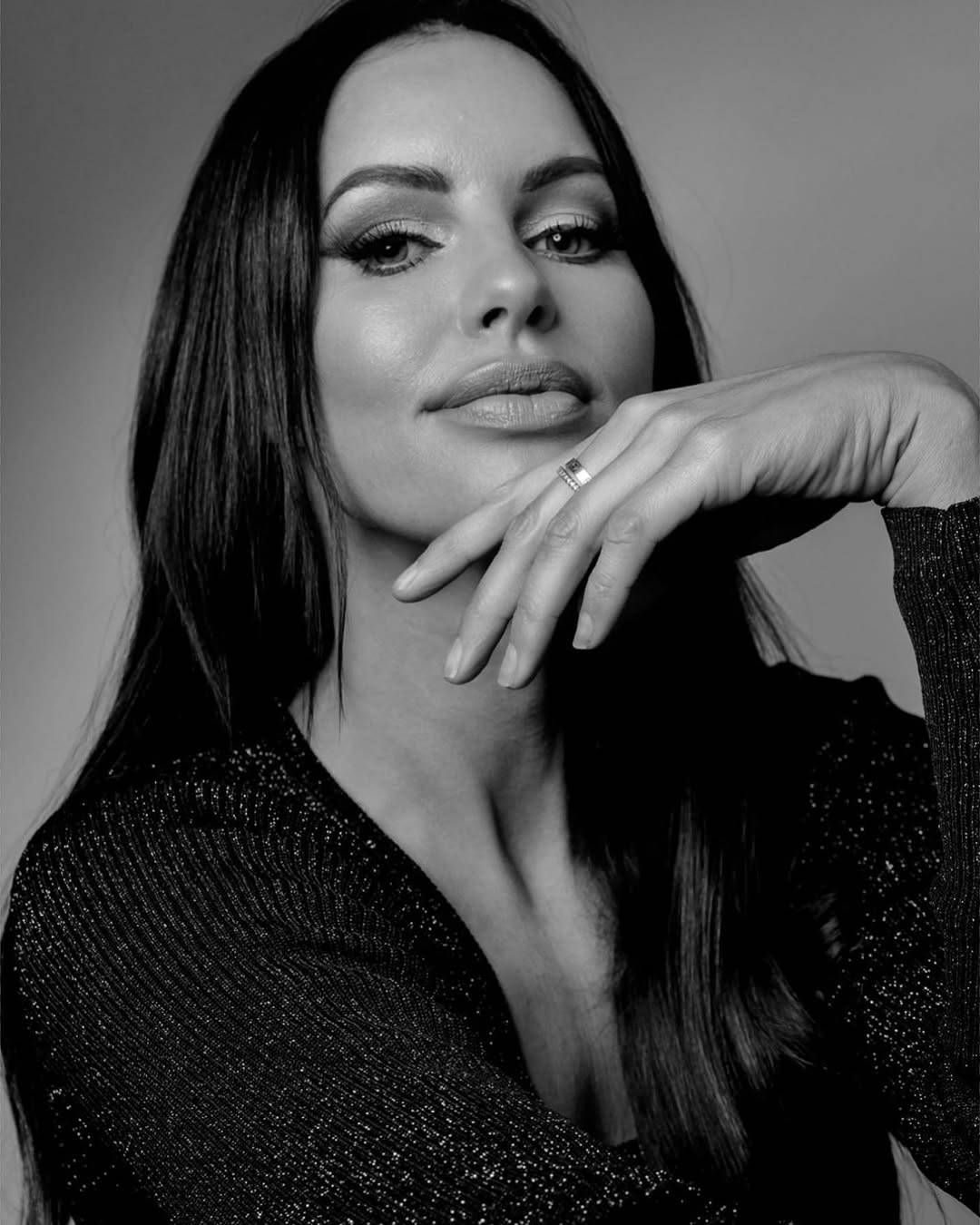 Striking black and white portrait of a brunette woman with hand on chin, wearing a shimmering top against a neutral backdrop.