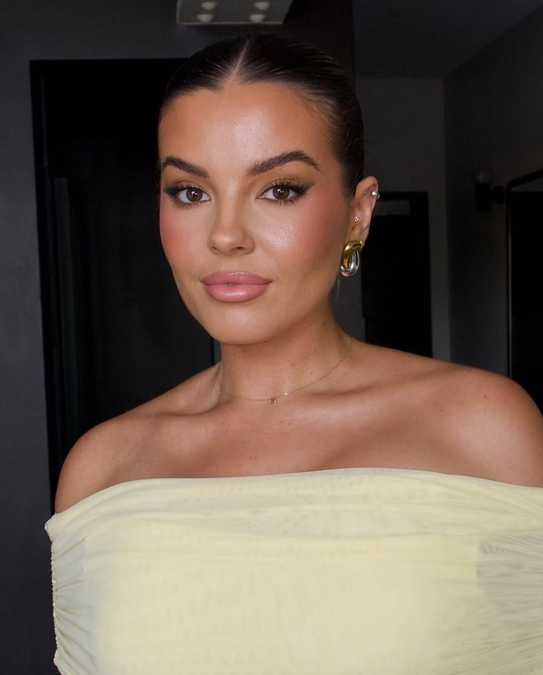 Close-up portrait of a woman with elegant makeup, accessorized with gold earrings and a necklace, wearing a light yellow off-shoulder dress.