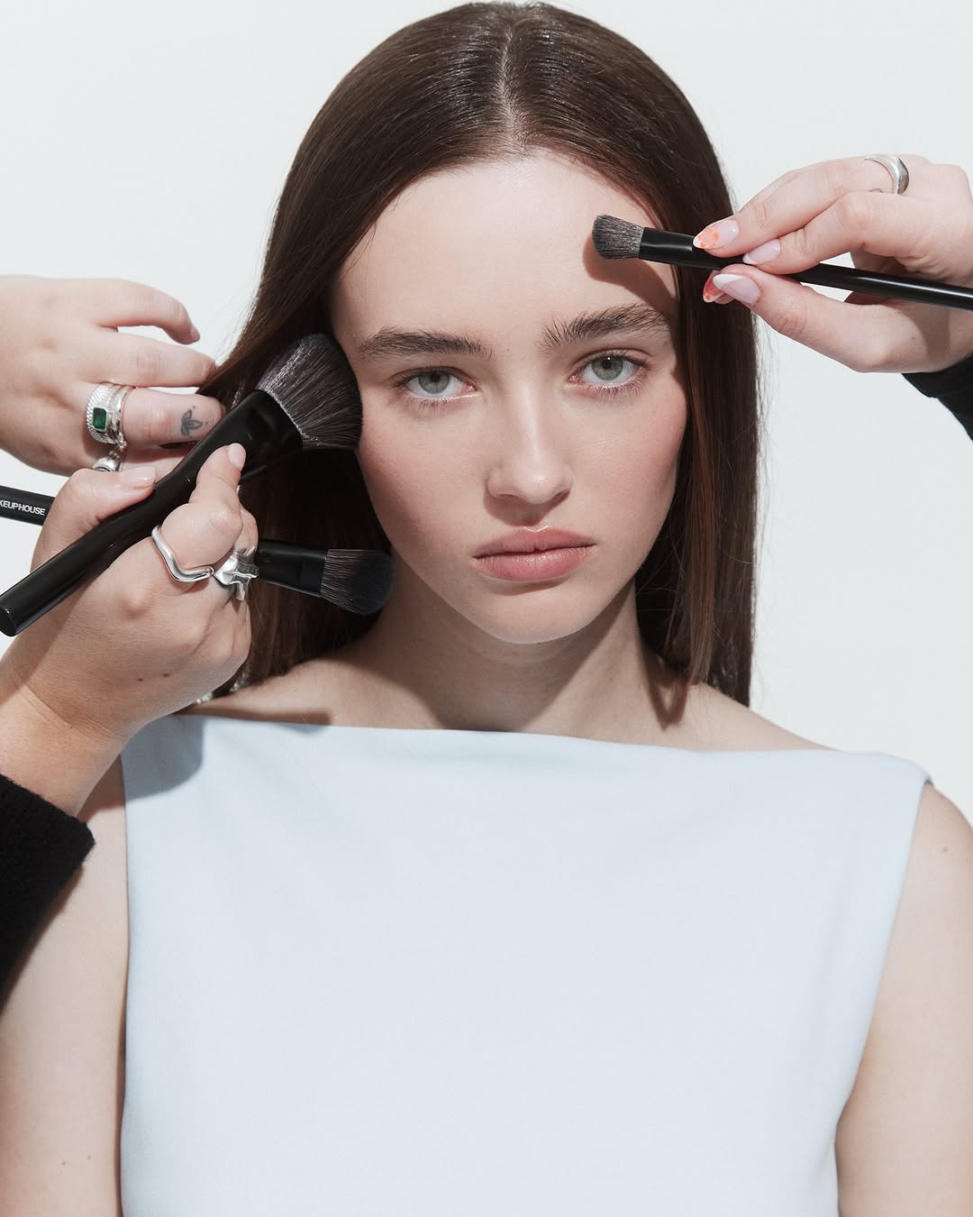 Portrait of a young woman having makeup applied with multiple brushes, set against a clean white background in a beauty shoot.