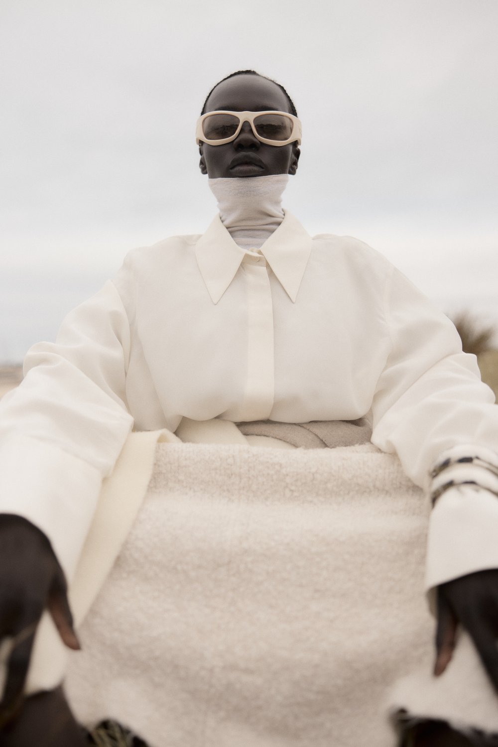 Elegant portrait of a Black woman in an all-white outfit with glasses, a turtleneck, and a collared shirt against a neutral backdrop.