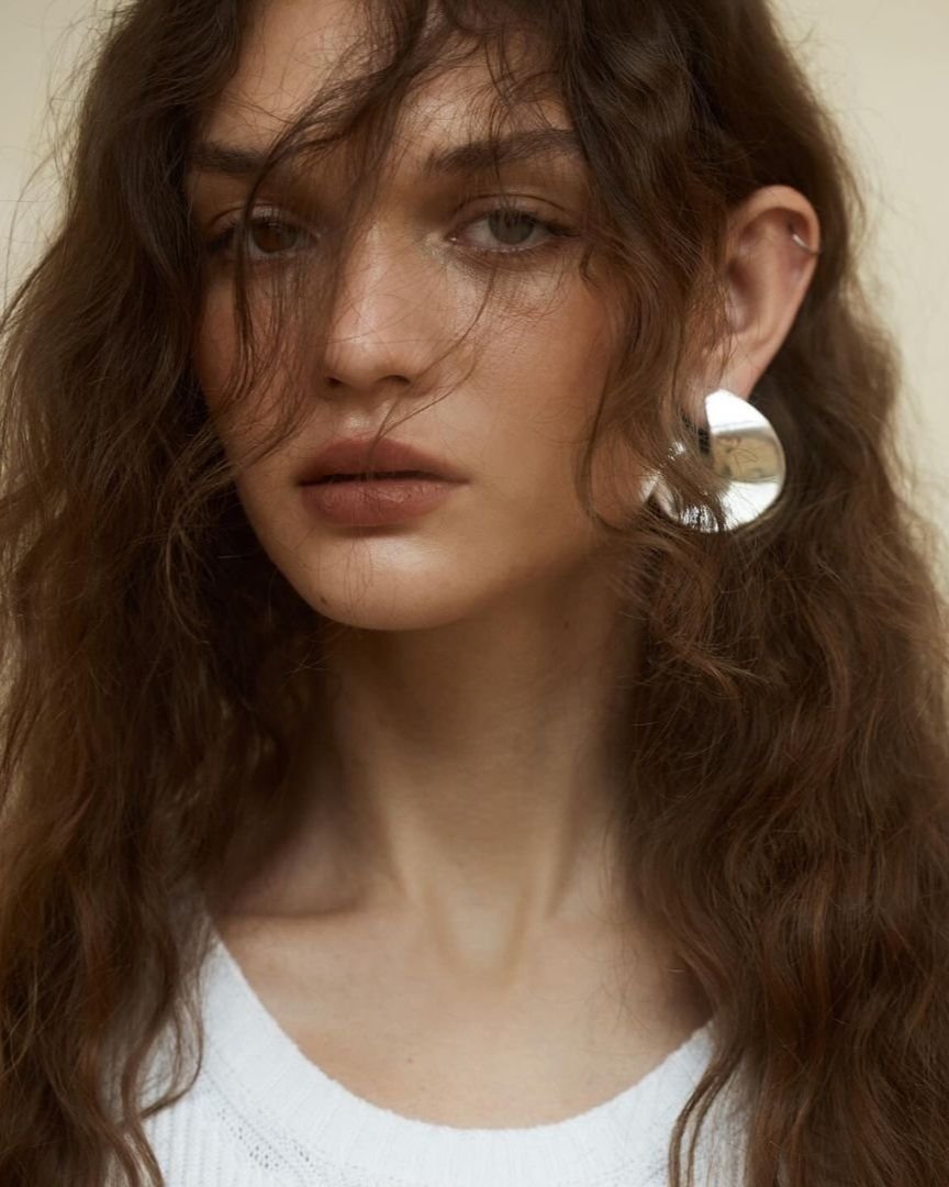 Close-up of an adult female with curly brown hair, wearing a white shirt and silver earrings, posing indoors with a neutral expression.