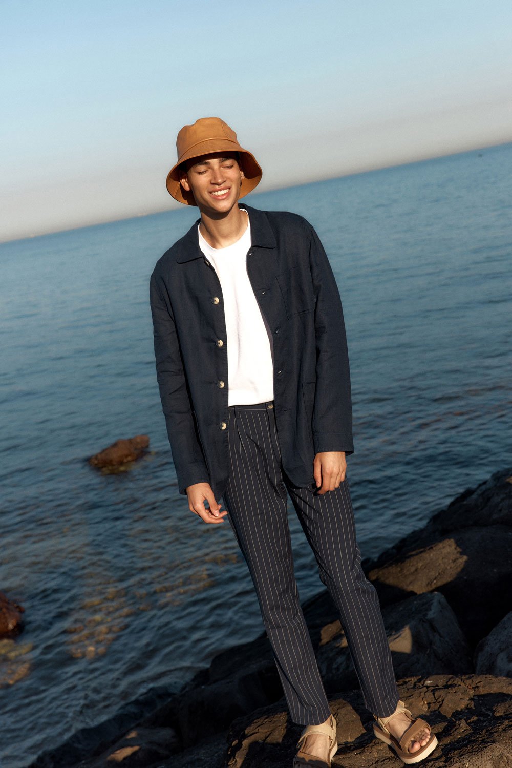 Young man in a blue jacket, striped pants, and hat poses on rocks by the sea on a sunny day. Fashion shot with natural light.