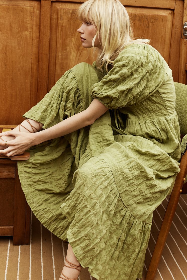 Blonde woman poses in a green, textured dress with tie-up sandals, seated against a light brown paneled wall; fashion or lifestyle shoot.