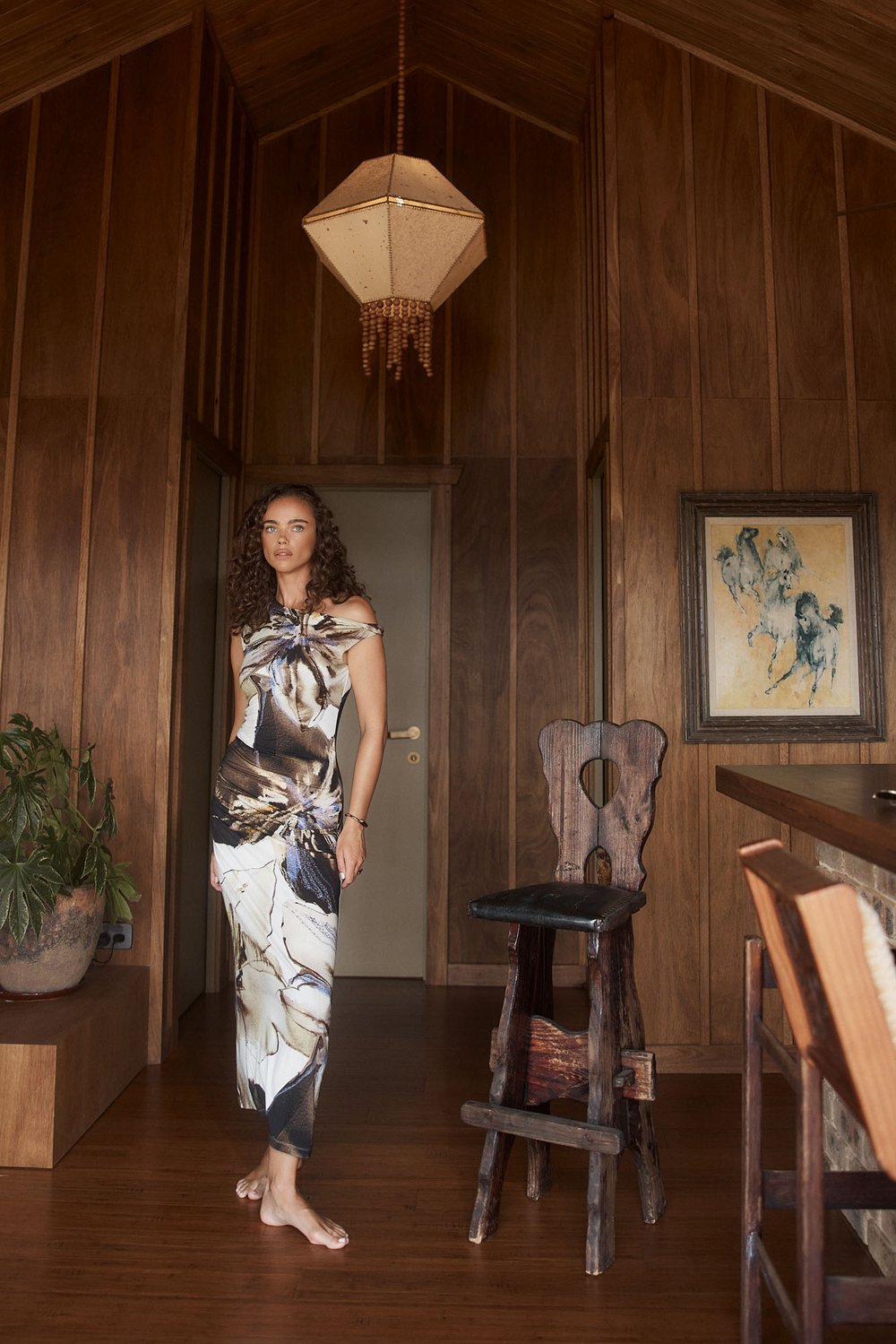 Adult woman in a floral dress stands barefoot in a wood-paneled home interior with vintage decor, bathed in soft, natural lighting.