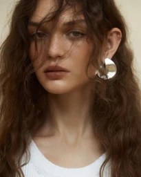 Close-up portrait of a woman with long wavy brown hair wearing a white top and a large silver earring