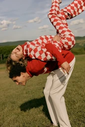 Man giving a piggyback ride to a smiling woman wearing red and white checkered trousers in a grassy field outdoors.