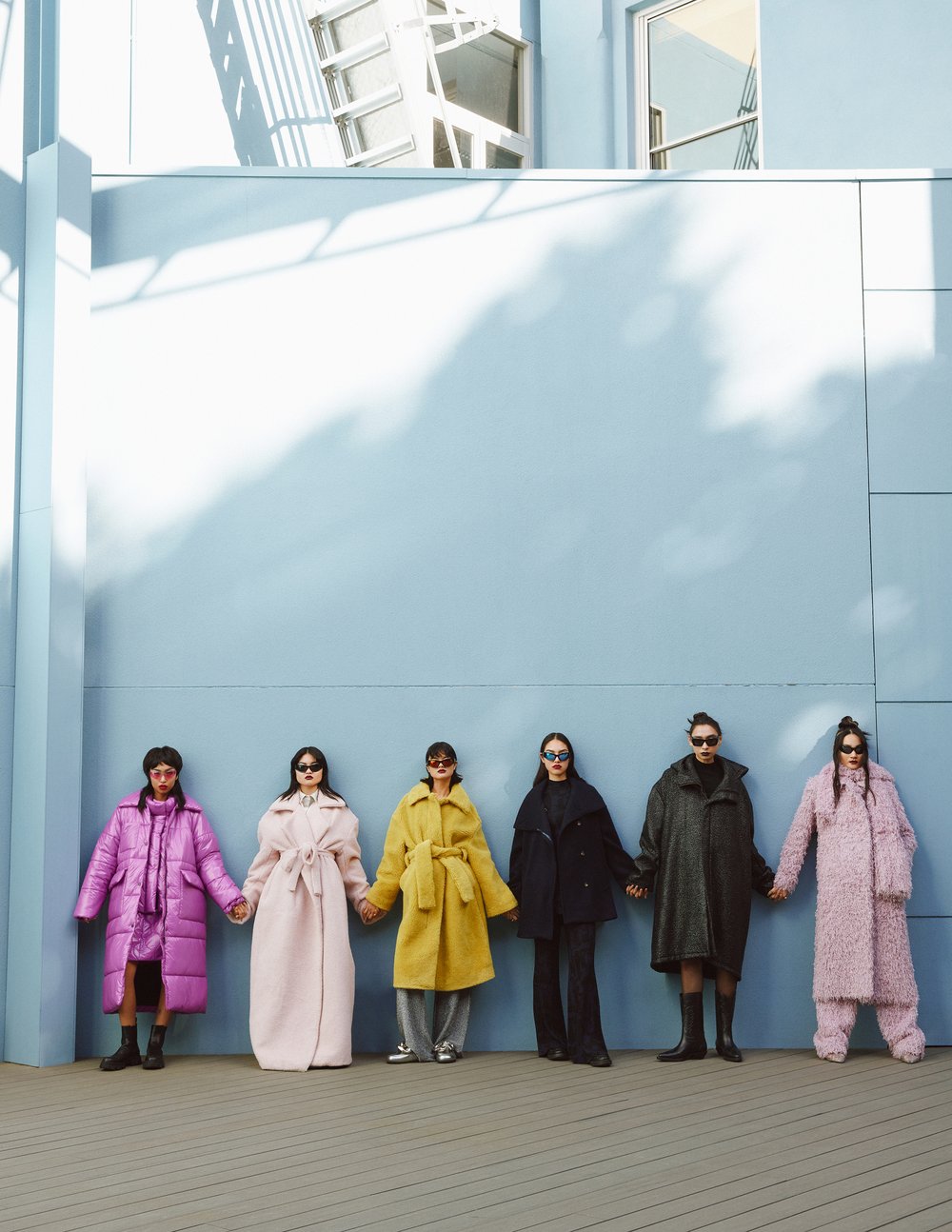 Six women wearing colorful winter coats holding hands in front of a light blue wall and grey wood floor in an outdoor setting.