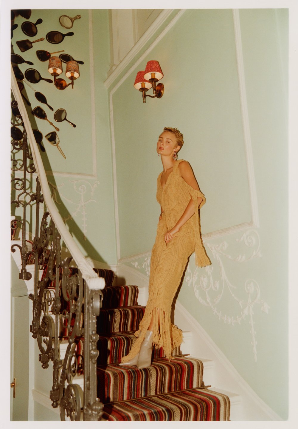 Woman in a tan dress and boots descends a staircase inside a vintage-style home with patterned carpet and decorative wall art and sconces.