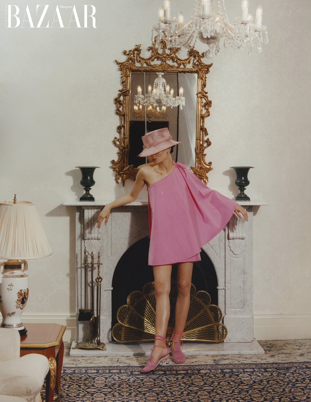 A woman in a pink dress and hat stands in front of a marble fireplace and ornate mirror in a high-fashion editorial shot indoors.