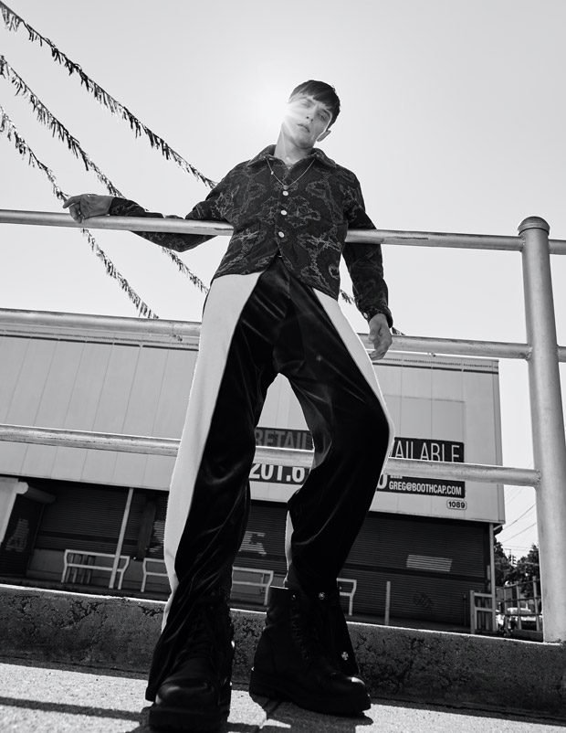 Monochrome fashion shot of a male model in patterned shirt and track pants leaning on a railing outdoors with building in the background.