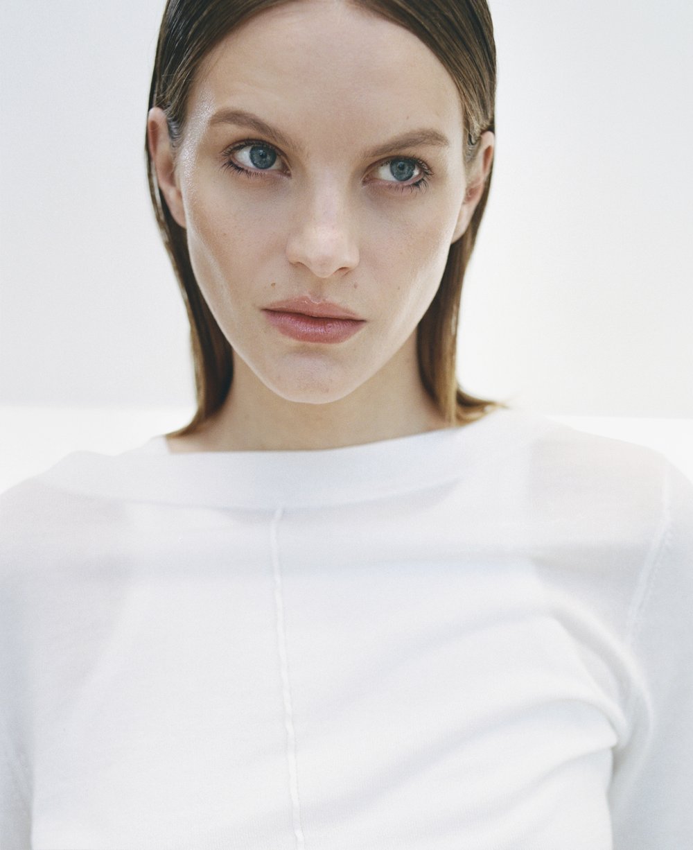 Close-up studio portrait of a young adult woman with blue eyes and brown hair, wearing a white top against a neutral background.