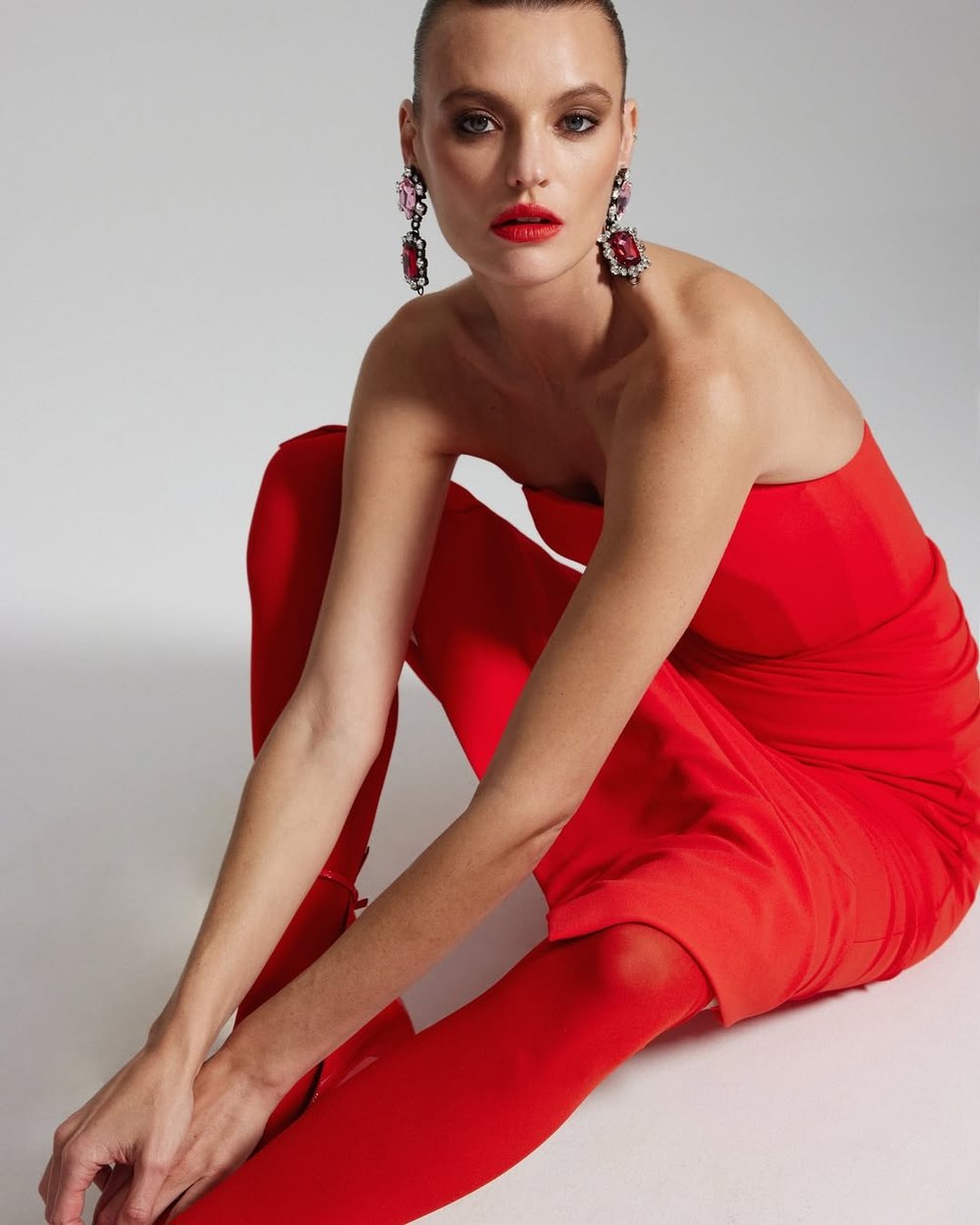 Elegant woman with red lipstick and matching outfit poses gracefully, accessorized with statement earrings, against a light background.