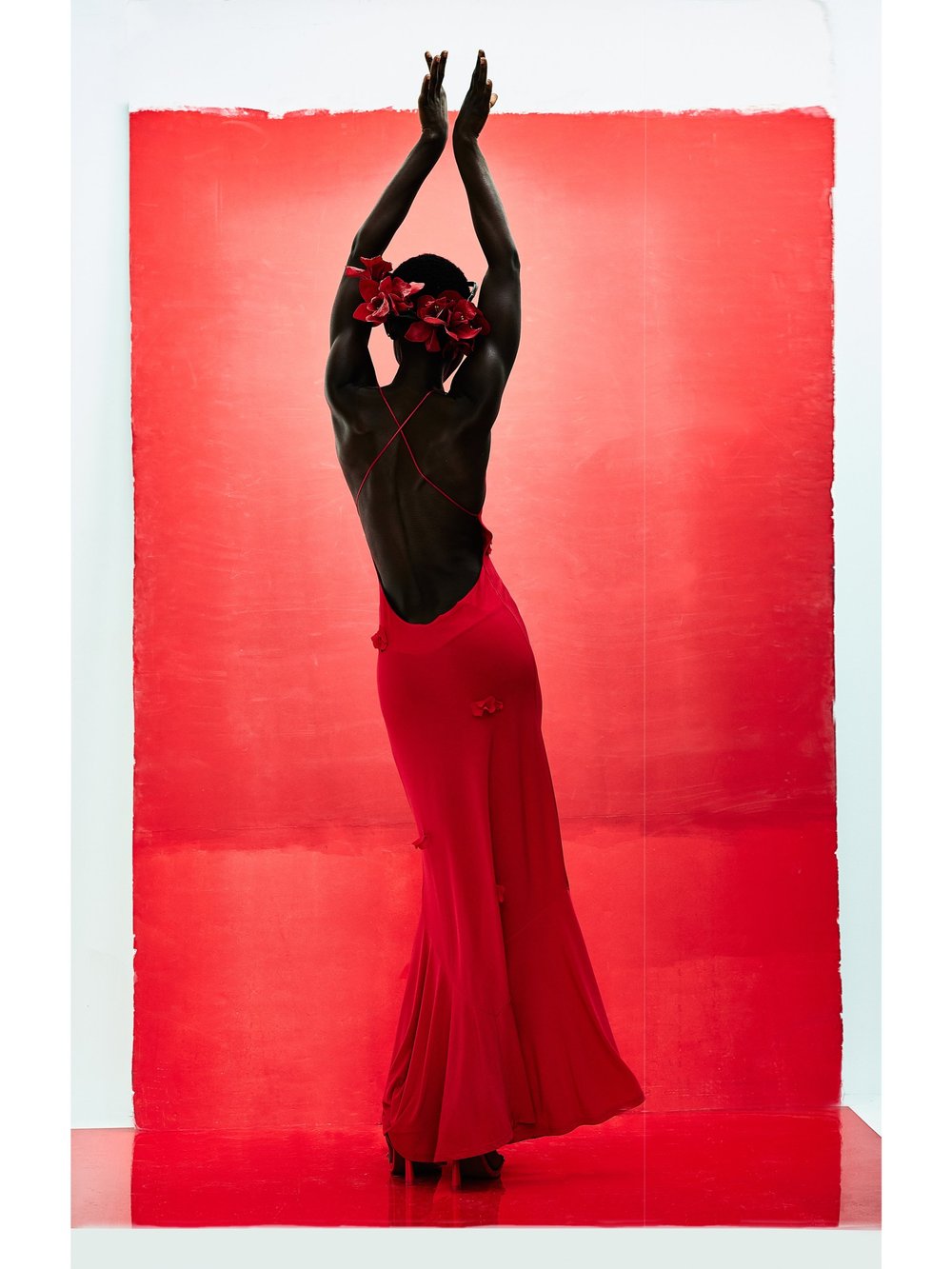 Woman in a red gown poses with arms raised against a red backdrop, wearing floral headpiece, in a fashion or dance-themed editorial.
