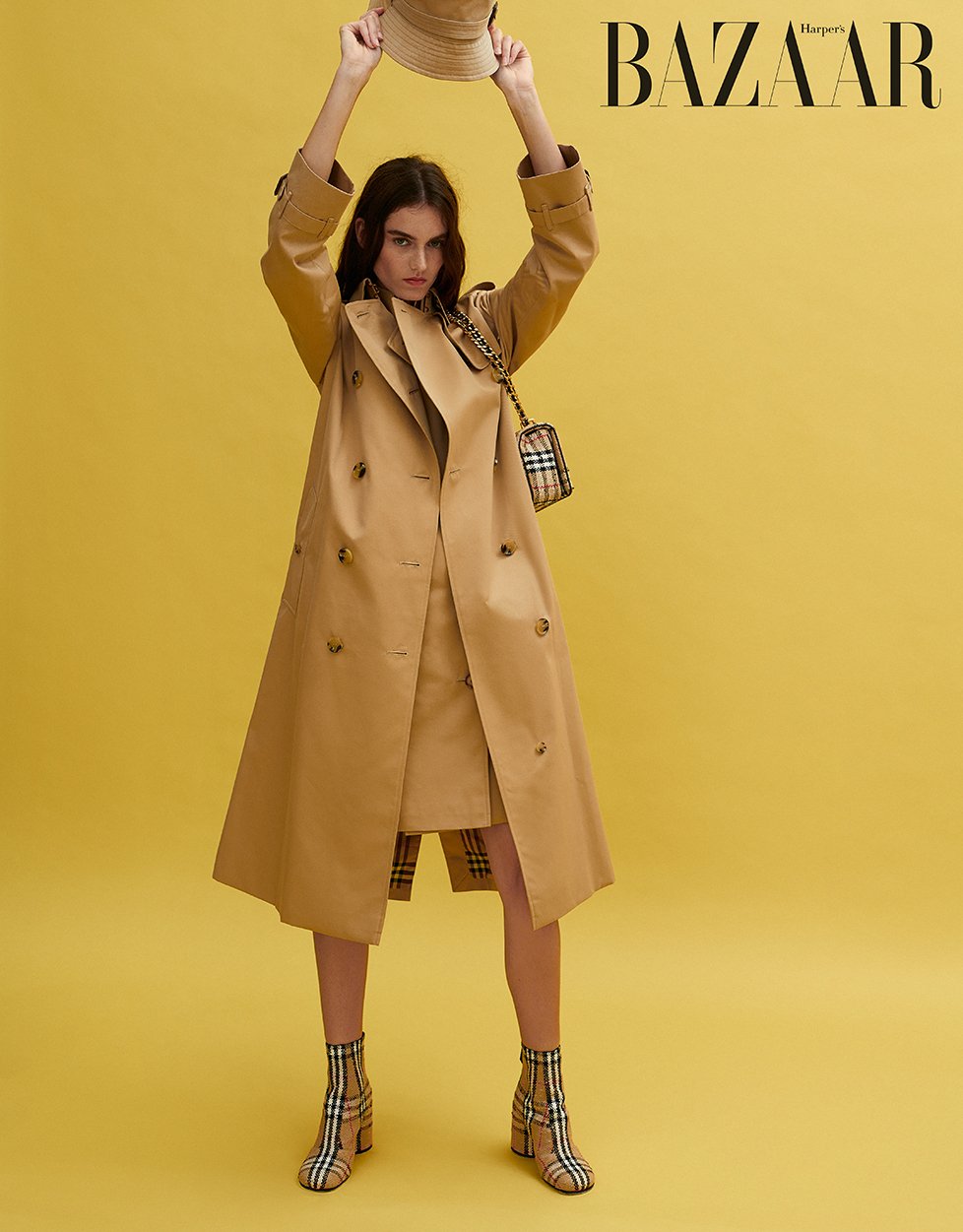 Fashion editorial shot of a woman in tan trench coat and matching skirt, holding a hat, against a yellow background with 'BAZAAR' text.