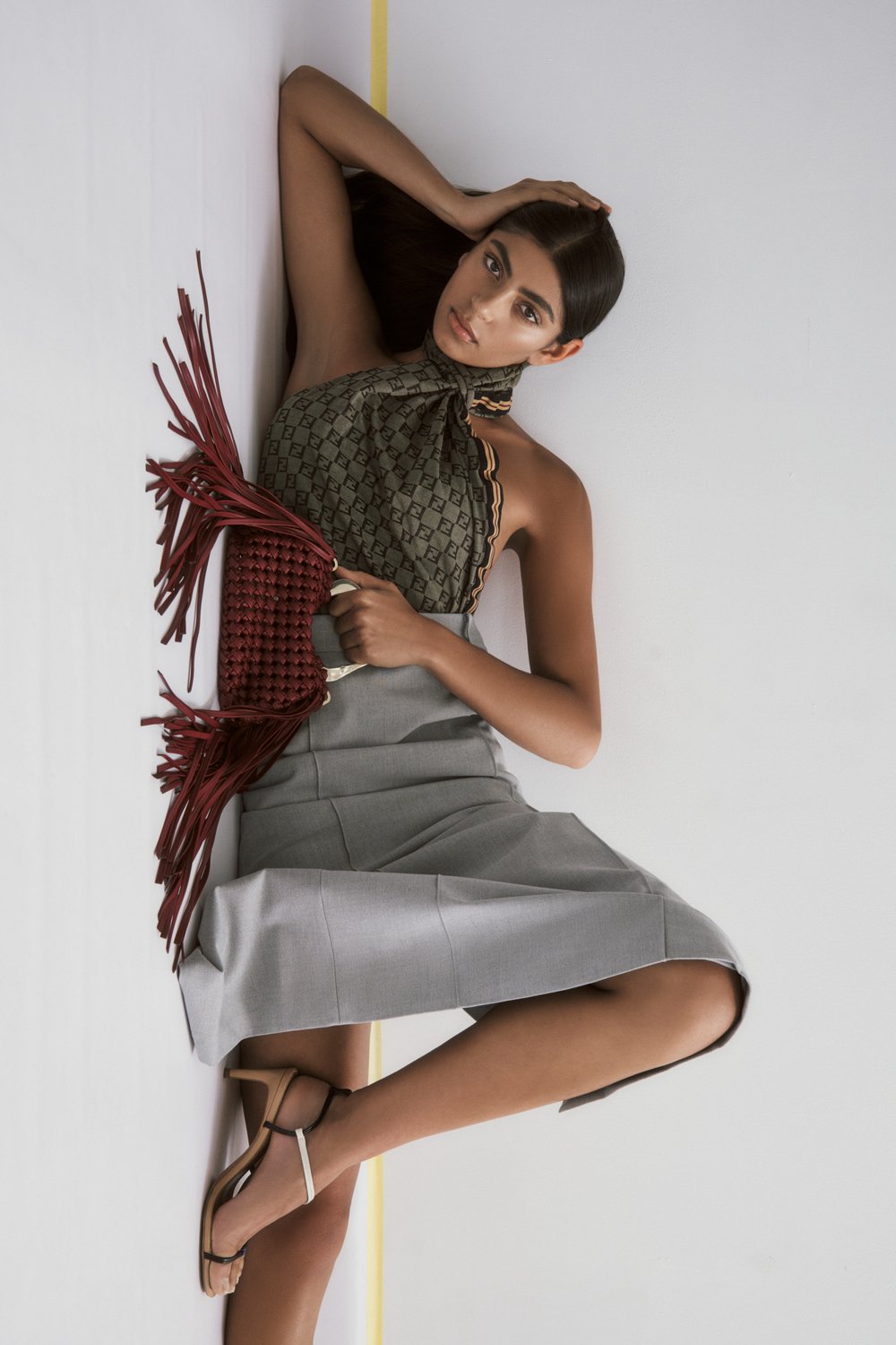 Fashion shot of a young woman lying near a wall wearing a printed top, grey skirt, and sandals while holding a fringe bag.