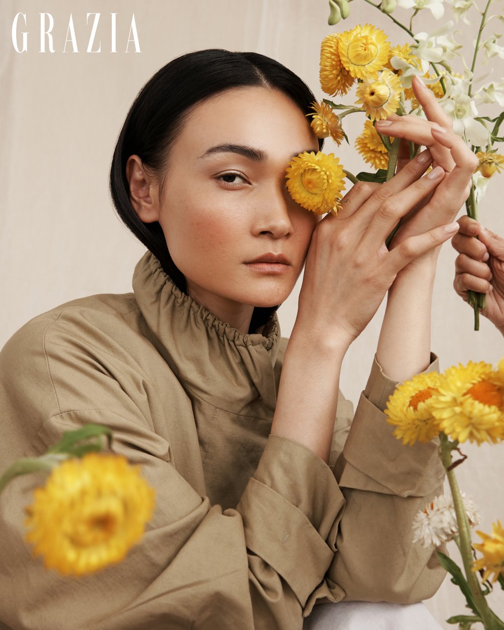 Fashion portrait of a young Asian woman with flowers covering one eye, wearing a beige jacket, set against a cream backdrop.