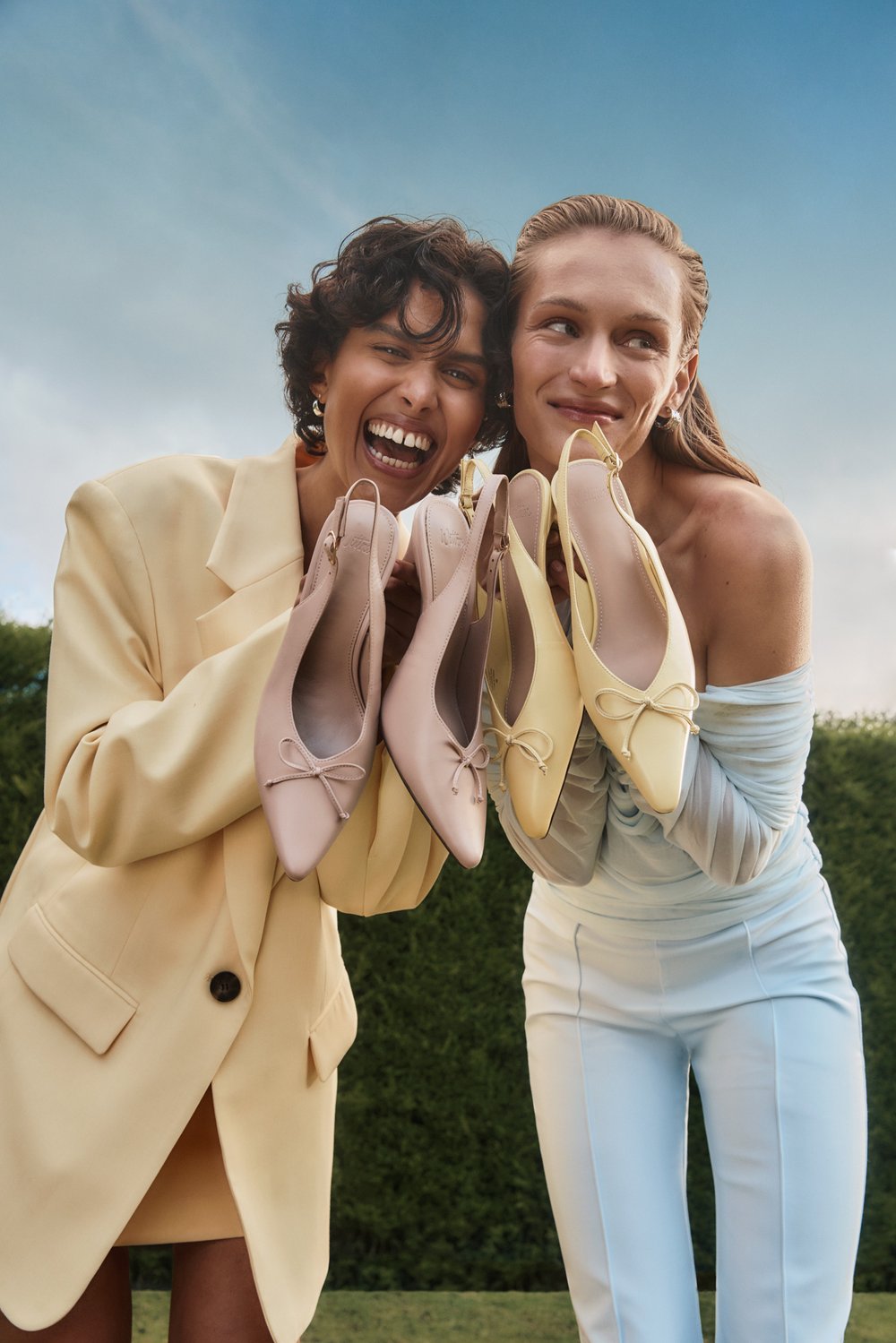 Two models display pastel-colored ballet flats outdoors against a blurred sky, one smiling broadly and wearing a yellow blazer.