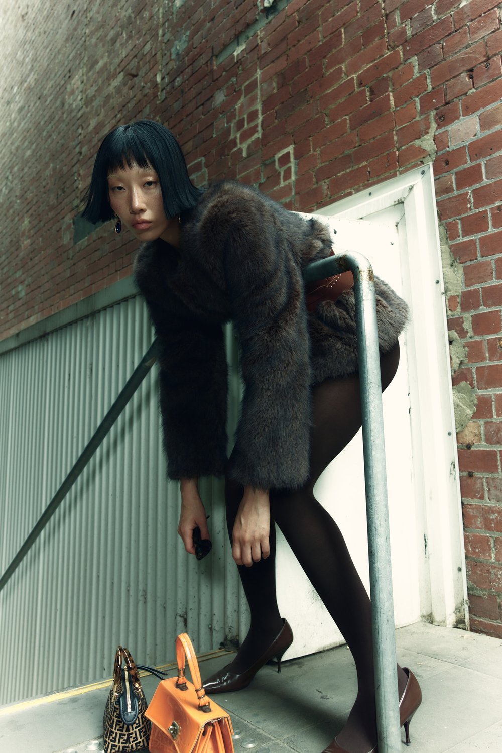 Brunette woman in a fur jacket poses by a metal railing near a brick wall with purses on the ground in an urban setting.